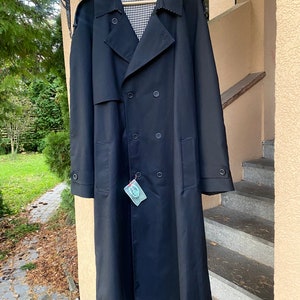 Op de afbeelding: Een lange zwarte trenchcoat met een dubbele rij knopen en een geruite voering. De jas heeft een label eraan.