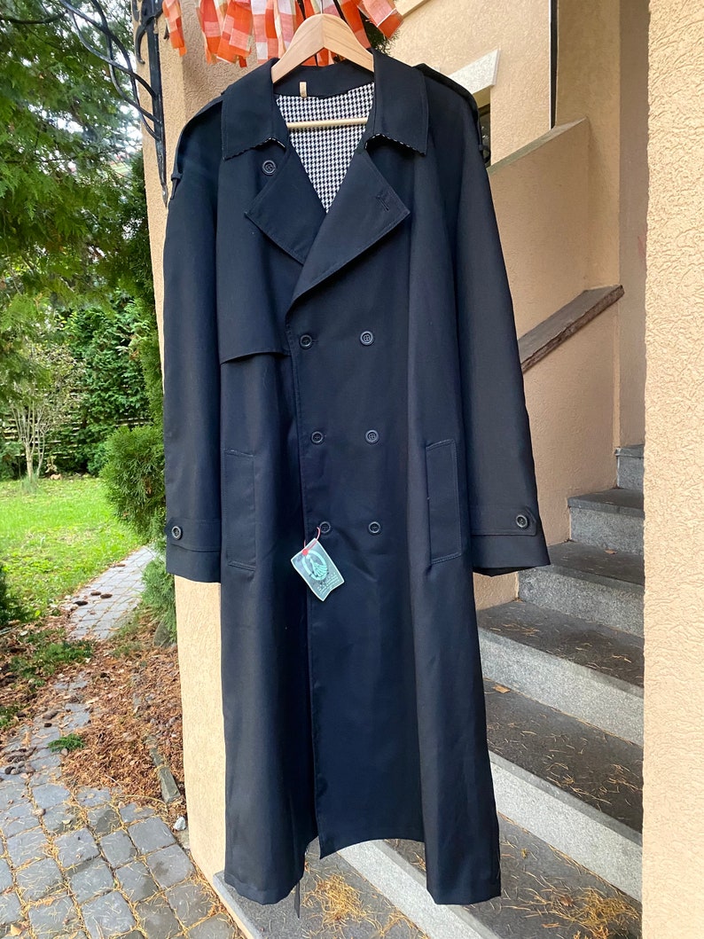Op de afbeelding: Een lange zwarte trenchcoat met een dubbele rij knopen en een geruite voering. De jas heeft een label eraan.