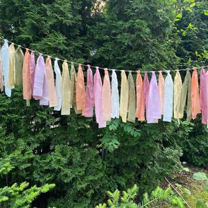 Könnte beinhalten: Eine Girlande aus bunten Stoffstreifen hängt vor einem Hintergrund aus grünem Laub. Die Streifen sind in verschiedenen Pastelltönen gehalten, darunter Rosa, Pfirsich, Gelb und Weiß. Das dekorative Banner ist an einer dünnen Schnur aufgereiht.