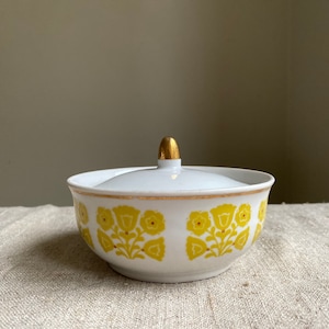 Könnte beinhalten: Eine weiße Zuckerdose aus Keramik mit Deckel und goldener Verzierung. Die Dose ist mit einem gelben Blumenmuster verziert.