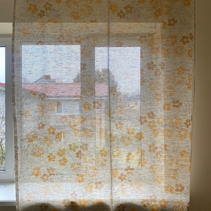 May include: A pair of sheer white curtains with a yellow floral pattern. The curtains are hanging in a window and the light is shining through them.