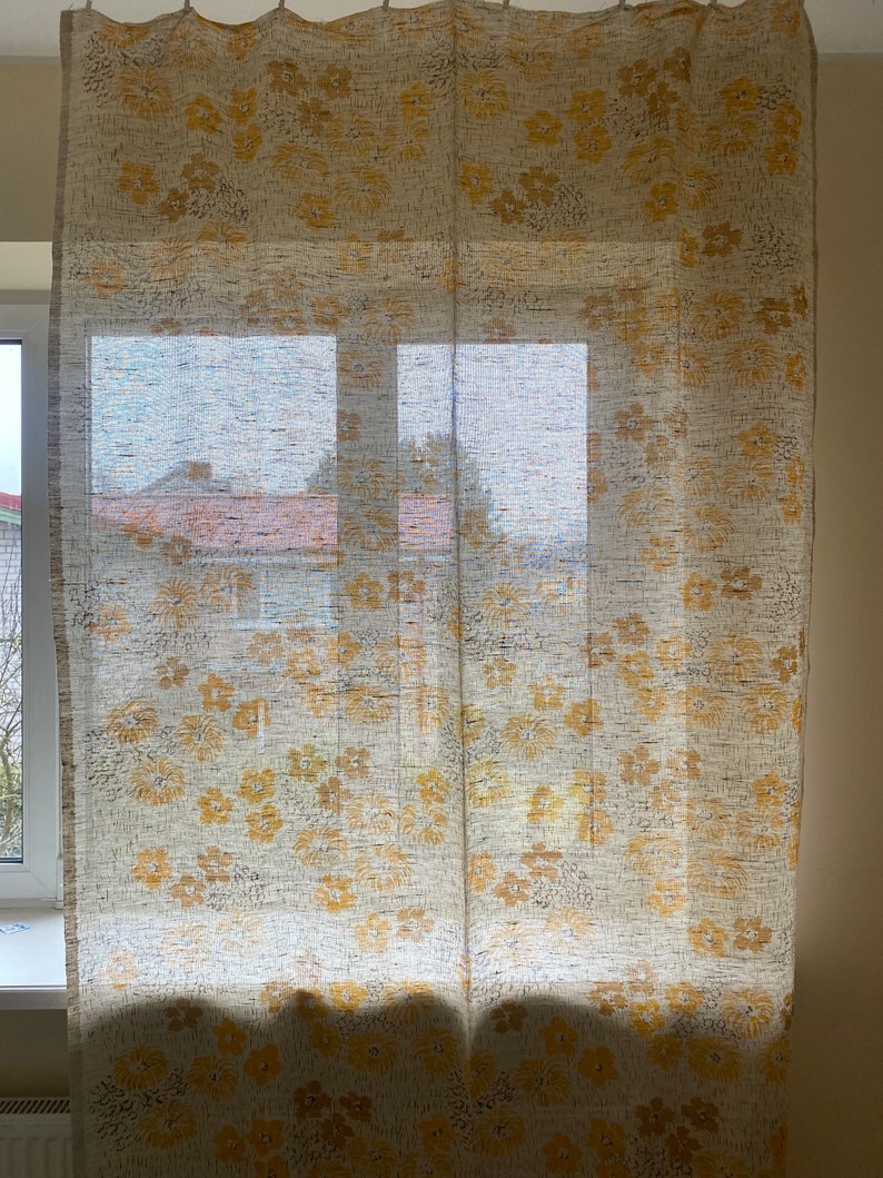 May include: A pair of sheer white curtains with a yellow floral pattern. The curtains are hanging in a window and the light is shining through them.