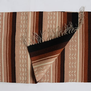 May include: A woven rug with a striped pattern in shades of brown, pink, and white. The rug has a fringe border and is folded in half.
