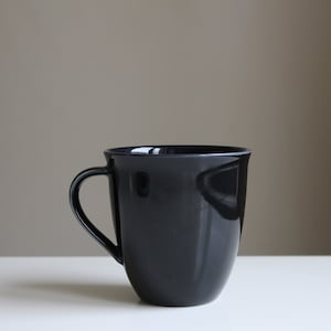 Puede incluir: Una taza de cer&aacute;mica negra con forma redondeada y asa. La taza est&aacute; sobre una superficie blanca.