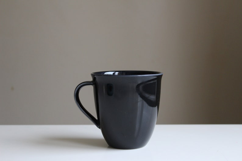 Puede incluir: Una taza de cer&aacute;mica negra con forma redondeada y asa. La taza est&aacute; sobre una superficie blanca.
