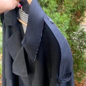 Op de afbeelding: Een zwarte trenchcoat met een zwart-wit geruite voering. De jas heeft een dubbele rij knopen en een grote kraag.