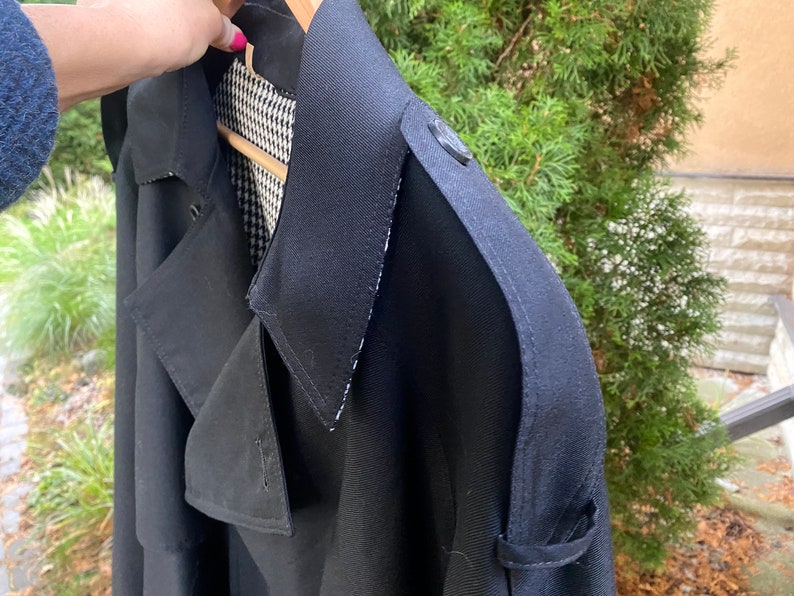 Op de afbeelding: Een zwarte trenchcoat met een zwart-wit geruite voering. De jas heeft een dubbele rij knopen en een grote kraag.