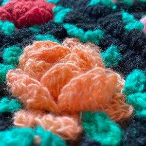 May include: Close-up of a crocheted fabric with a pink flower and green leaves. The fabric is black and has a textured surface.