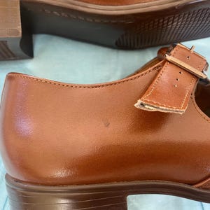 May include: A pair of brown leather shoes with a buckle strap. The shoes have a low heel and a rounded toe.