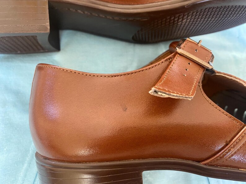 May include: A pair of brown leather shoes with a buckle strap. The shoes have a low heel and a rounded toe.