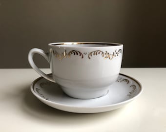 May include: A white teacup and saucer with a gold trim and floral design.