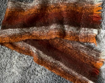 Op de afbeelding: Een pluizige, getextureerde sjaal met horizontale strepen in tinten van gebrand oranje, donkerbruin en crème. De sjaal heeft een zachte, geborstelde uitstraling met gerafelde, gefranjerde randen aan de uiteinden en een warm kleurenpalet.