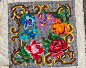 May include: A vintage floral embroidery on a cream-colored fabric with a gray background. The embroidery features a variety of flowers in shades of pink, red, blue, yellow, and green, surrounded by a gold-colored frame.