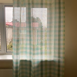May include: A white window with a green and white checkered curtain. The curtain is sheer and allows light to pass through. The window is closed and the curtain is pulled to the side.