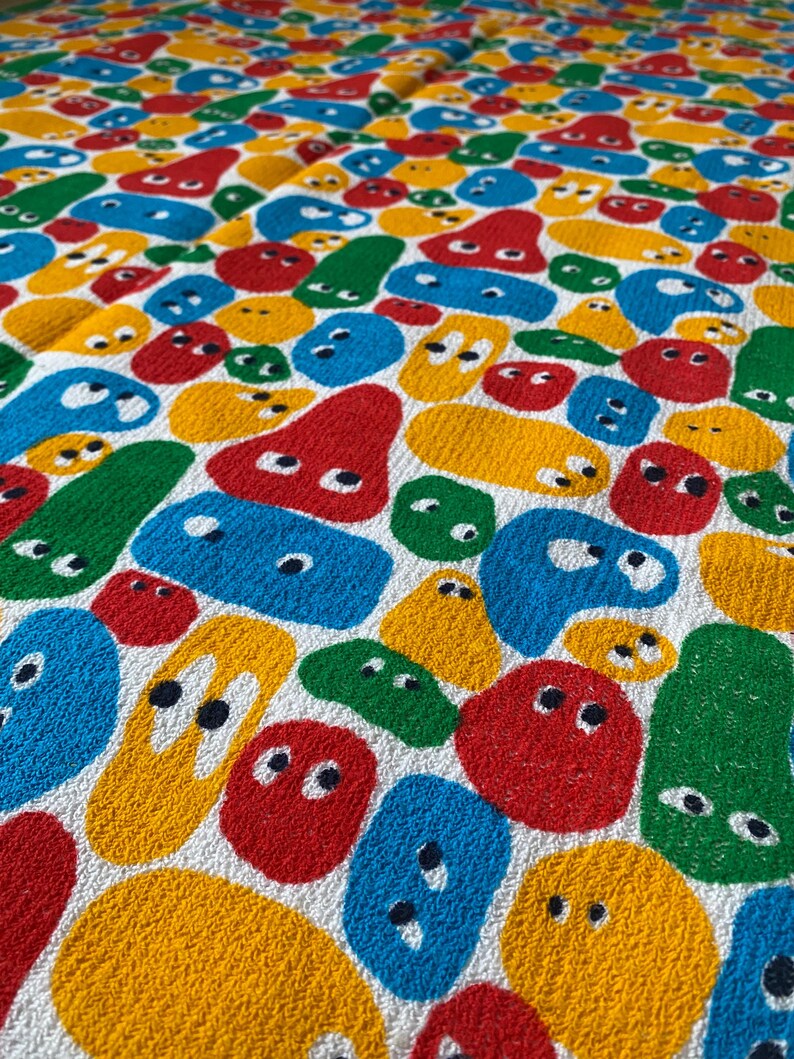 Pu&ograve; includere: Un tessuto bianco con un motivo ripetuto di forme astratte colorate. Le forme sono rosse, gialle, blu e verdi, ognuna con due piccoli punti neri che assomigliano a occhi. Il design &egrave; giocoso e vibrante.