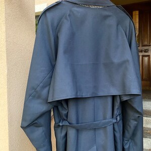 Op de afbeelding: Een donkerblauwe trenchcoat met een grote kraag en een riem. De jas heeft een dubbele rij knopen en een enkele knoop bovenaan.