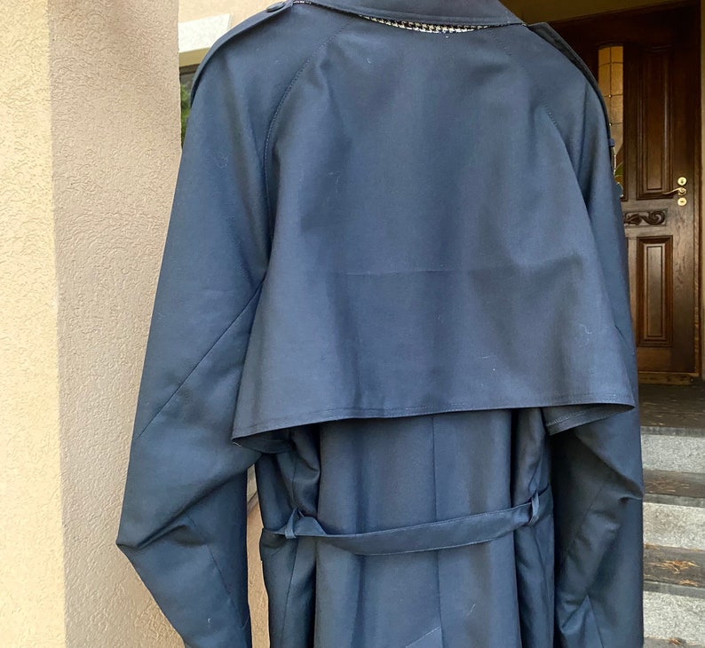 Op de afbeelding: Een donkerblauwe trenchcoat met een grote kraag en een riem. De jas heeft een dubbele rij knopen en een enkele knoop bovenaan.