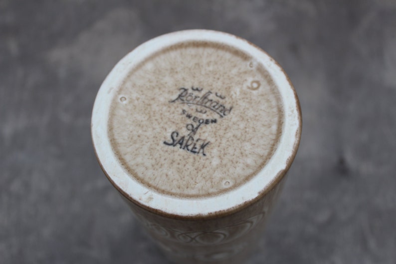 K&ouml;nnte beinhalten: Der Boden einer wei&szlig;en Keramikvase mit brauner Glasur. Die Vase ist mit dem Text "R&ouml;rstrand Sweden" und "SAREK" gekennzeichnet.