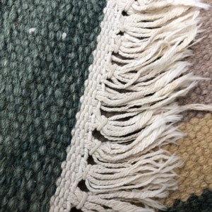 May include: Close-up of a woven rug with a fringe border. The rug has a green, brown, and beige color scheme.