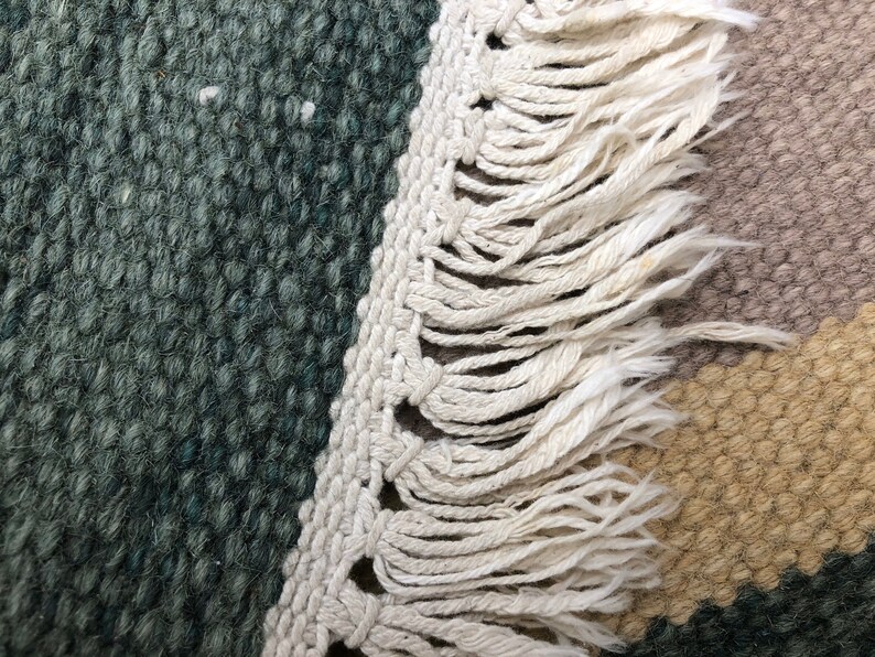May include: Close-up of a woven rug with a fringe border. The rug has a green, brown, and beige color scheme.