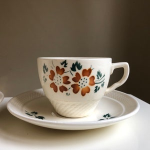 Peut inclure: Une tasse &agrave; th&eacute; et une soucoupe blanches avec un motif floral dans les tons de brun et de vert. La tasse a une forme arrondie et une anse. La soucoupe a un bord festonn&eacute;.
