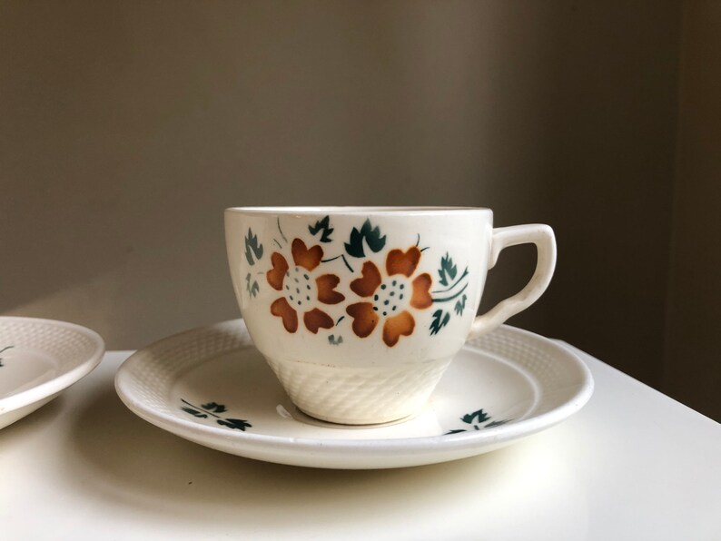 Peut inclure: Une tasse &agrave; th&eacute; et une soucoupe blanches avec un motif floral dans les tons de brun et de vert. La tasse a une forme arrondie et une anse. La soucoupe a un bord festonn&eacute;.