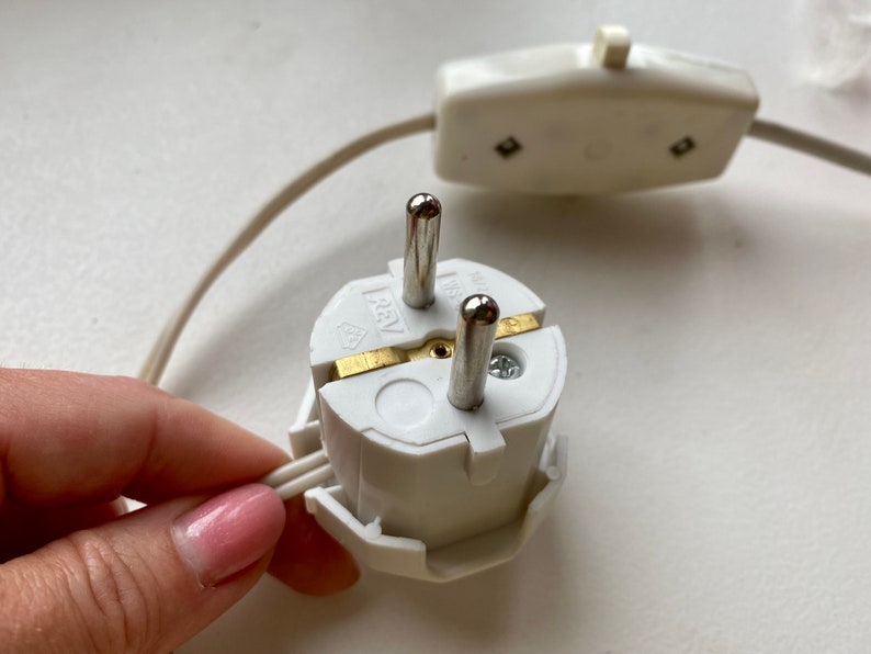 Może przedstawiać: Biała wtyczka elektryczna z dwoma bolcami i bolcem uziemiającym. Wtyczka jest podłączona do białego przewodu z włącznikiem w linii.
