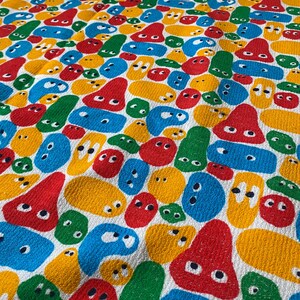 Pu&ograve; includere: Un tessuto colorato con un motivo ripetuto di forme astratte in rosso, giallo, blu e verde. Le forme hanno occhi bianchi e sono su uno sfondo bianco. Il tessuto ha un aspetto strutturato.