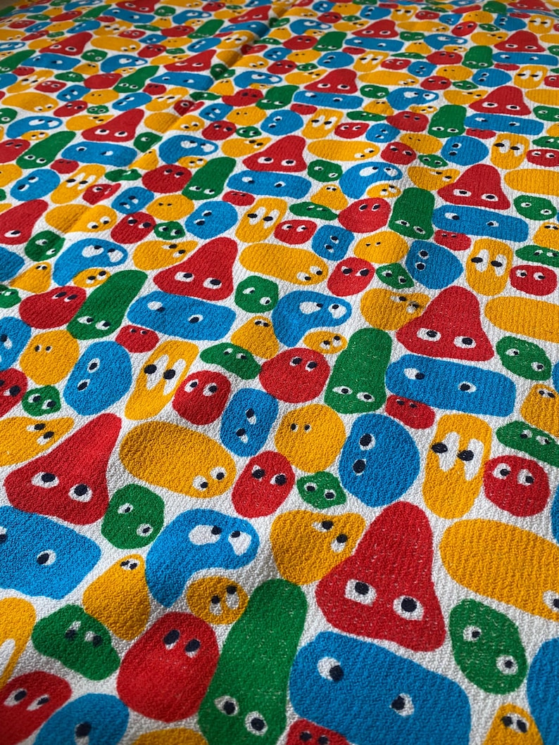 Pu&ograve; includere: Un tessuto colorato con un motivo ripetuto di forme astratte in rosso, giallo, blu e verde. Le forme hanno occhi bianchi e sono su uno sfondo bianco. Il tessuto ha un aspetto strutturato.