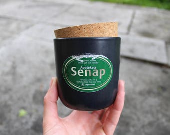 May include: A black jar with a cork lid and a green label that says "Apotekets Senap" with the text "Förvaras kallt, 250 g, bäst före 09-2003, Apoteket".