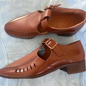 May include: A pair of brown leather shoes with cutouts and buckles. The shoes have a textured pattern and are on a light blue surface.