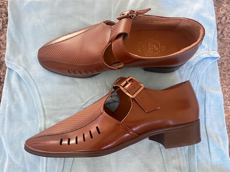 May include: A pair of brown leather shoes with cutouts and buckles. The shoes have a textured pattern and are on a light blue surface.