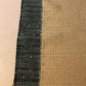 May include: Close-up of a woven rug with a beige, dark brown, and dark blue color scheme. The rug has a textured surface and a fringe border.