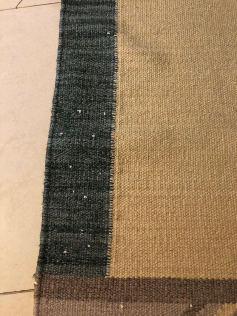 May include: Close-up of a woven rug with a beige, dark brown, and dark blue color scheme. The rug has a textured surface and a fringe border.
