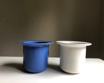 May include: Two ceramic planters, one blue and one white. Both planters have a wide rim and a cylindrical shape.
