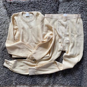 May include: Cream-colored thermal underwear set, consisting of a long-sleeved crew neck top and matching long johns. The waffle-knit fabric has a ribbed texture. The set is laid out on a gray, textured surface.