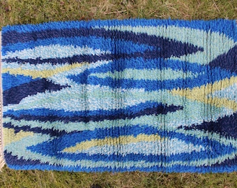 May include: A blue, green, and yellow abstract patterned rug with fringed edges. The rug has a modern, geometric design.