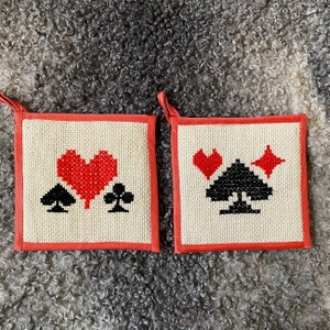 Puede incluir: Dos agarraderas rojas y blancas con símbolos de corazón y cartas de juego. Las agarraderas están hechas de tela blanca con un borde rojo.