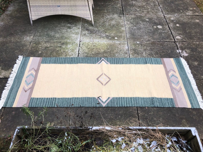 May include: A long, rectangular rug with a cream background and a geometric pattern in shades of green, pink, and blue. The rug has fringe on the ends.
