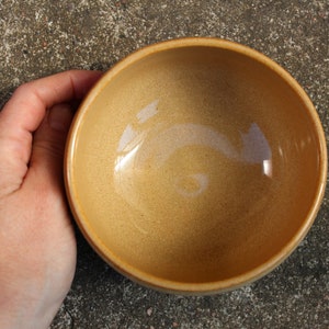 May include: A light brown ceramic bowl with a smooth, slightly textured surface. The bowl is empty and appears to be made of stoneware.