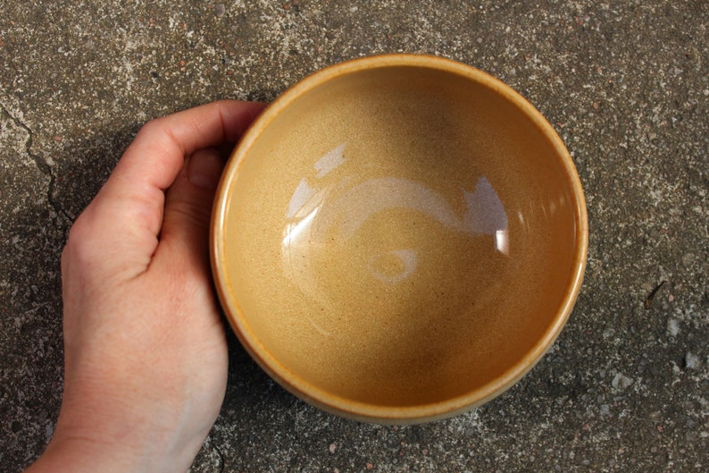 May include: A light brown ceramic bowl with a smooth, slightly textured surface. The bowl is empty and appears to be made of stoneware.