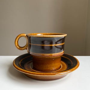 Puede incluir: Un juego de taza y platillo de café de cerámica marrón. La taza tiene una banda marrón oscuro con una raya marrón más clara. El platillo tiene un diseño a juego. El juego está sobre una superficie blanca.