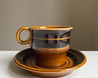 Puede incluir: Un juego de taza y platillo de café de cerámica marrón. La taza tiene una banda marrón oscuro con una raya marrón más clara. El platillo tiene un diseño a juego. El juego está sobre una superficie blanca.
