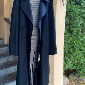 Op de afbeelding: Een lange, donkerblauwe trenchcoat met een zwart-wit geruite voering. De jas heeft een dubbele rij knopen, een riem en twee zakken.