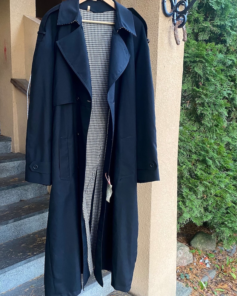 Op de afbeelding: Een lange, donkerblauwe trenchcoat met een zwart-wit geruite voering. De jas heeft een dubbele rij knopen, een riem en twee zakken.