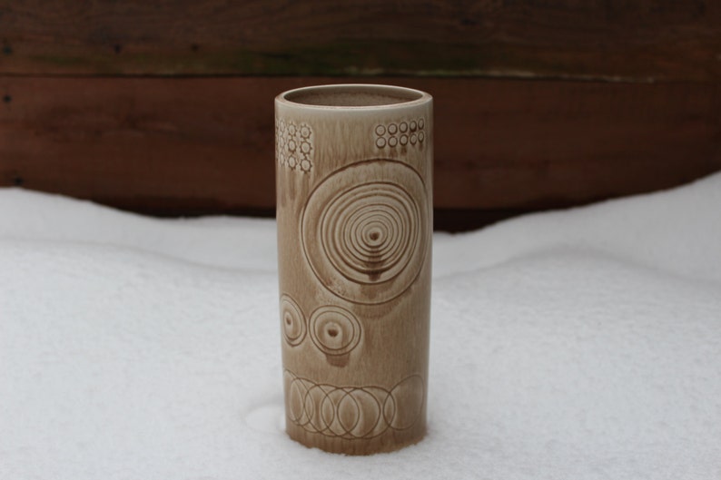 K&ouml;nnte beinhalten: Eine beige Keramikvase mit einem geometrischen Muster aus Kreisen und Ovalen. Die Vase steht auf einer wei&szlig;en Oberfl&auml;che.
