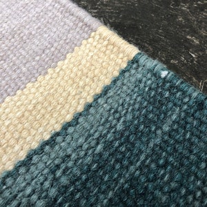 May include: Close-up of a woven rug with three horizontal stripes in shades of light purple, beige, and teal.