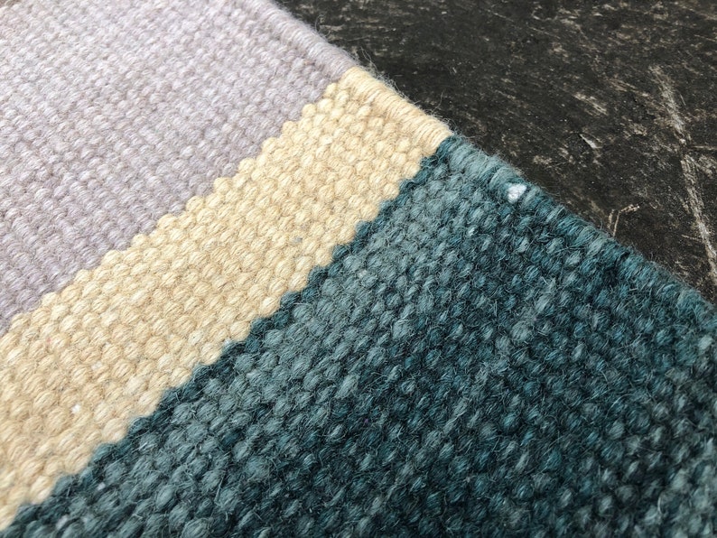 May include: Close-up of a woven rug with three horizontal stripes in shades of light purple, beige, and teal.