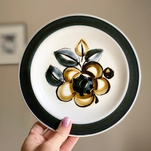 Puede incluir: Un plato de cerámica blanco con un borde verde y un diseño floral. La flor es amarilla con detalles marrones y negros.