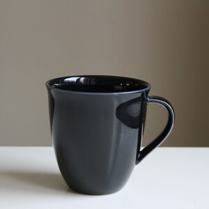 Puede incluir: Una taza de cer&aacute;mica negra con forma redondeada y asa.